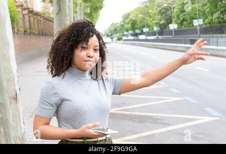 Ein junges schwarzes Mädchen mit lockigen Haaren in der Stadt, das ein Taxi hochhagelt, während es ein Handy in der Hand hält. Stockfoto