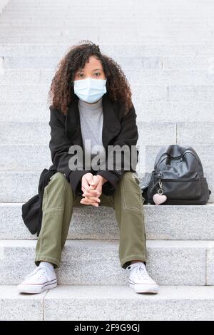 Junge Frau mit Maske sitzt auf der Treppe und ruht ihre Arme auf den Knien, wobei ihre Hände zusammengeklemmt sind und direkt auf die Kamera blicken. Stockfoto