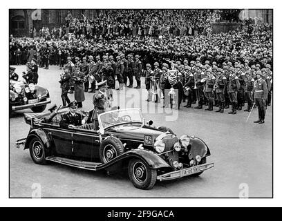 Adolf Hitler in Uniform, der in seinem offiziellen offenen Mercedes-Wagen mit Hakenkreuzarmband steht, grüßt das Militär und die Massen bei einer großen Nazi-Kundgebung im Jahr 1938 in Deutschland. Hitler begrüßt die Teilnehmer eines Reichsparteitages in Nürnberg. Ein uniformierte Martin Bormann und Dr. Goebbels saßen ebenfalls im Heck eines offenen Mercedes-Autos Stockfoto