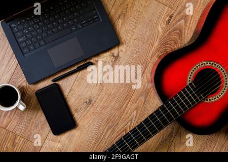 Rot-schwarze klassische Gitarre auf Holzboden, neben Computer, Telefon und Notebook-Stift Stockfoto