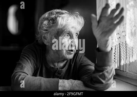 Die alte Frau spricht gestikulierend am Tisch in ihrem Haus. Schwarzweiß-Foto. Stockfoto