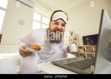 Fröhlicher, fetter Mann im Schlafanzug, der morgens auf dem Bett saß, das Frühstück genoss und lachte Stockfoto