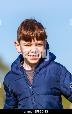 Ein süßer, blauäugiger, rothaariger Junge in einer blauen Jacke, der an einem sonnigen Frühlingstag auf einem Park läuft Stockfoto