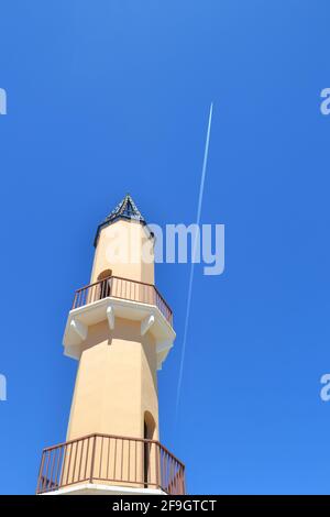 Orange-gelber Turm in der Marina von Fuengirola, gegen den perfekten klaren blauen Himmel, als das Flugzeug über dem Vapor Trail fliegt - Fuengirola, Andalusien.