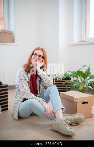 Eine hübsche junge Frau mit Brille ist müde, als sie in eine neue Wohnung umzieht, sie sitzt auf dem Boden neben Kisten und einer Topfpflanze und träumt davon. Stockfoto