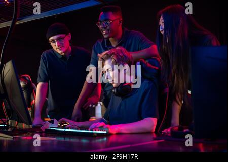 Zwei Jungs und ein Mädchen in medizinischen Masken in einem Computerclub unterstützen ihren Freund während seines Spiels. Stockfoto