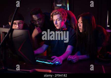 Zwei Jungs und ein Mädchen in medizinischen Masken in einem Computerclub unterstützen ihren Freund während seines Spiels. Stockfoto