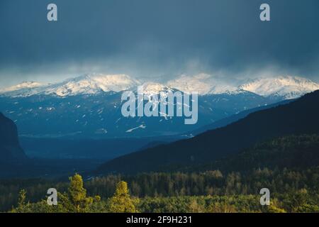 Sonnenbeschienene Bergkette mit Schneespitzen und immergrüner Waldlandschaft bei stürmischem Wetter im Inntal, Tirol, Österreich Stockfoto