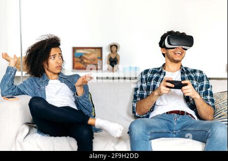 Afroamerikanisches Mädchen und gemischter Renner verbringen Freizeit zusammen zu Hause auf einem Sofa. Aufgeregt Kerl verwendet Virtual-Reality-Brille für Videospiele, ein gelangweiltes Mädchen sitzt neben ihm und sieht ihn empört Stockfoto