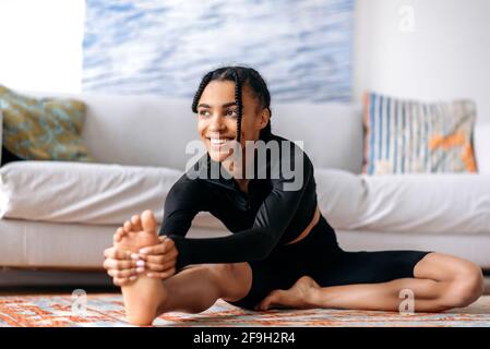 Sportlich und flexibel hübsches afroamerikanisches Mädchen in Sportkleidung, engagiert sich für Fitness, führt einen gesunden Lebensstil, dehnt sich zu Hause auf dem Boden, schaut zur Seite, lächelt fröhlich Stockfoto