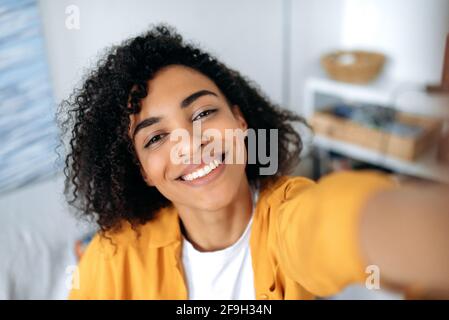 Selfie-Foto auf dem Smartphone. Fröhlich sorglos lockiges afroamerikanisches Mädchen, Schülerin, Freiberuflerin, macht ein Selfie am Telefon, täuscht sich herum, hat Spaß, schaut auf die Webcam des Telefons, lächelt glücklich Stockfoto