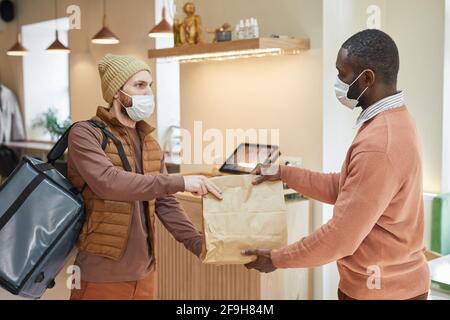 Seitenansicht Porträt eines afroamerikanischen Mannes, der im Café den Auftrag an den auslieferenden Mann übergibt, beide mit Masken, covid Konzept, Kopierraum Stockfoto