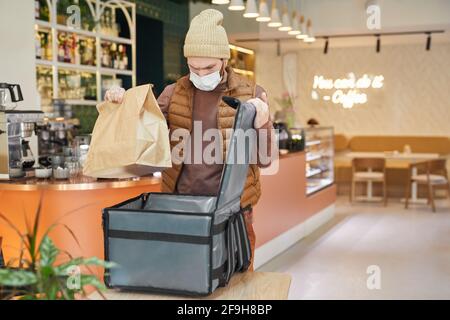 Taille hoch Porträt der Lieferung Mann trägt Maske beim Verpacken Aufträge in Café-Innenraum zu Tasche, kopieren Raum Stockfoto