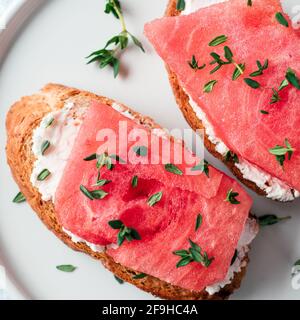 Toast mit Frischkäse und Wassermelone. Salzige käse, süßes Wassermelone und würziger Thymian auf knusprig gegrillte Brotscheiben. Idee und Rezept ungewöhnlich gesundes Frühstück, Snack Sommer. Top Aussicht, flach. Platz kopieren Stockfoto