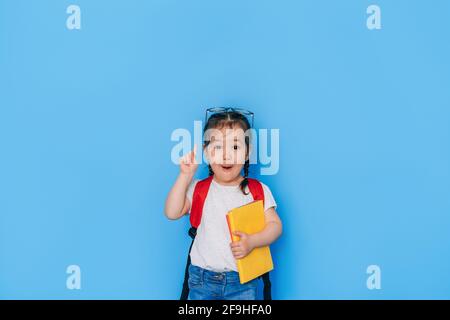 Überrascht schulmädchen hält gelbe Buch vor blauem Hintergrund. Zurück zur Schule im Jahr 2021. Homeschooling, Fernunterricht. Stockfoto