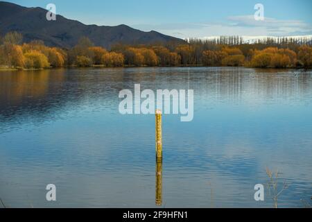 Wasserstand Messmast Hintergrund sind Berge und Bäume im Fall eines Sees auf der ländlichen Südinsel, Neuseeland. Stockfoto