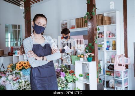 Frau Managerin trägt Gesichtsmaske Floristin stehend gekreuzten Arm mit ihrem Personal im Hintergrund. Stockfoto