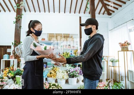 Lächelnd junge Kauffrau trägt eine Gesichtsmaske und Schürze. Wartung männliche Flanell Blume Käufer Stockfoto