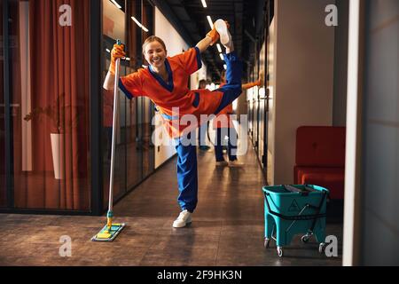 Fröhlicher Arbeiter, der eine einbeinige Haltung auf dem Mopp machte Stockfoto