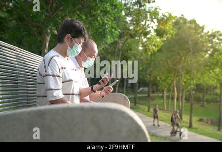 Ein asiatisches Ehepaar mit Gesichtsmaske sitzt und liest in einem Park ihre Telefone. Stockfoto