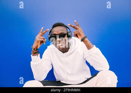 Junger afrikanischer schwarzer Mann saß an einer blauen Wand und schaute Bei der Kamera trägt ein weißes Sweatshirt und eine Sonnenbrille Siegeszeichen Stockfoto