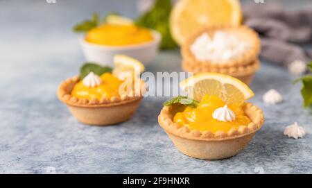 Minitörtchen mit Zitronenquark, Mini-Baiser, Zitronenscheiben und Minze auf blauem Betongrund. Selektiver Fokus. Stockfoto