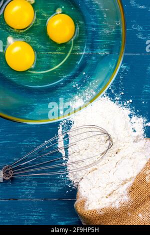 Zutaten für die Herstellung von Pfannkuchenteig. Huhn Eigelb in einer Glasschüssel, Hand Schneebesen und Mehl auf einem Holztisch. Stockfoto