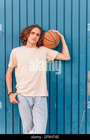 Vertikales Porträt eines jungen kaukasischen Mannes, der einen Basketball auf der Schulter hält. Er hat lange rote Haare und trägt lässige Sommerkleidung. Sie wird von einer blauen Metalltür getragen. Stockfoto