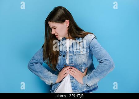 Kranke junge Frau legte die Hände auf den Bauch, der an Magenschmerzen litt und packte Bauchschmerzen fühlt sich schlecht zwielichtig an, gekleidet in Denim-Jacke Stockfoto