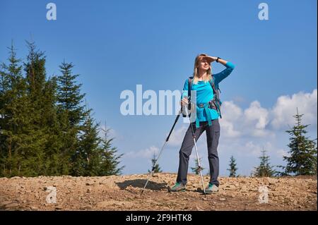 Weibliche Touristin, die auf einer schmutzigen Steinstraße steht und ihr Gesicht mit einer Hand bedeckt, die es vor hellem Sonnenlicht schützt, während sie ihren nächsten Weg beim Wandern beobachtet und studiert. Konzept des Reisens, Wanderns. Stockfoto