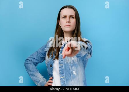 Porträt einer unglückten jungen Frau beschuldigt jemanden, zeigt direkt auf die Kamera, macht ernstes Gesicht, gekleidet in Jeansjacke, isoliert an blauer Wand Stockfoto