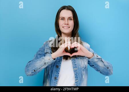 Fröhliche nette junge Frau mit breiten charmanten Lächeln legte Hände auf Brust machen mit Fingern Herzform Symbol der Nächstenliebe, isoliert auf blauen Studio Wand Stockfoto
