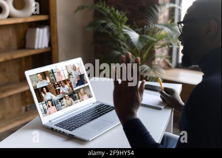 Seitenansicht lächelnder afrikanischer Geschäftsmann mit Brille winkend Hand, macht Videoanruf, grüßt Kollegen, Geschäftspartner diskutieren Projekt, mit Web-Kamera, virtuellen Chat, sitzen am Schreibtisch Stockfoto