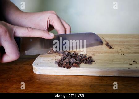 Ein Nahaufnahme-Bild einer Frau, die dunkle Stücke zerkleinern Schokolade in kleine Stücke mit einem Messer und einem Holz Schneidebrett, bevor es zu ihrem Herd hinzugefügt wird Stockfoto