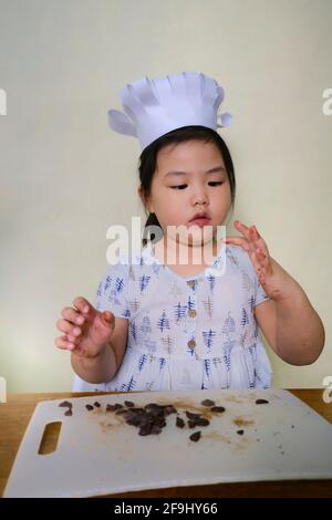 Ein süßes junges molliges asiatisches Mädchen mit einem weißen Kochmütze lernt mit ihrer Mutter zu kochen, Schokolade zu hacken und zu probieren, bevor es in den mischt Stockfoto