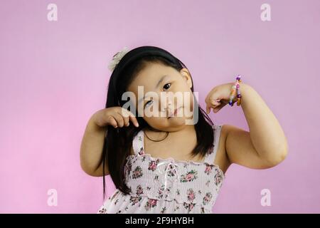 Ein süßes junges molliges asiatisches Mädchen posiert und zeigt ihren Bizeps, um zu zeigen, wie stark sie ist. Leuchtender rosafarbener Hintergrund. Stockfoto