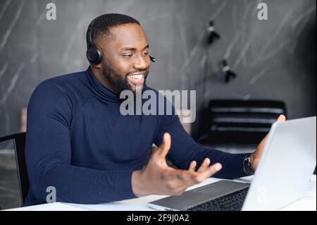 Fokussierter junger Geschäftsmann in einer Brille mit Kopfhörern, der Videoanruf mit Kunden auf dem Laptop hielt. Konzentrierter afrikanischer Mann in einer Brille, der Online-Unterricht hält und Kunden berät Stockfoto