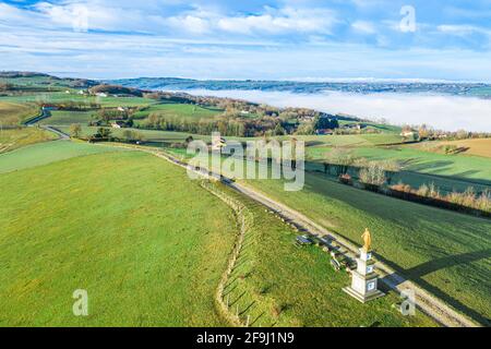 Drohnenaufnahme eines christlichen Denkmals und eines Pfades, der in eine kleine Stadt mit ausgedehnten, neblig-grünen Bergen im Hintergrund führt. Stockfoto