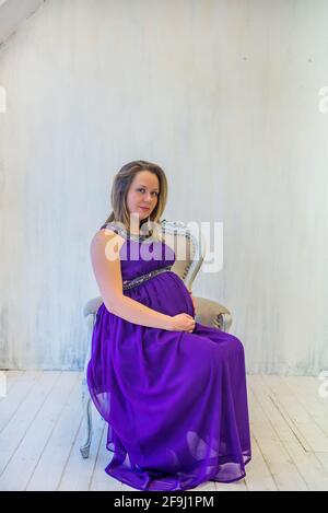 Schöne schwangere Frau in Ultra Violet Kleid sitzt auf dem Sessel Stockfoto