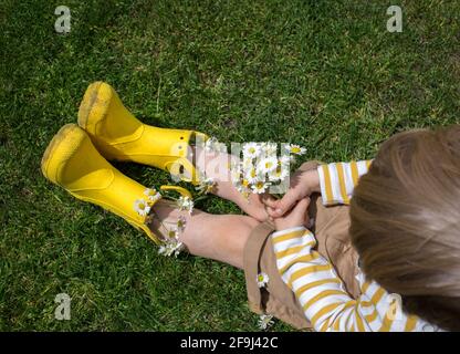 Unerkennbarer kleiner Junge in leuchtend gelben Gummistiefeln mit einem Strauß Gänseblümchen sitzt auf dem Gras. Blick von oben. Ein Geschenk mit Liebe für m gesammelt Stockfoto