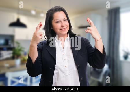 Lächelnde Frau Unternehmer trägt Büro smart casual Anzug machen doppelt Viel Glück Geste mit den Fingern kreuzte Home Room Hintergrund Stockfoto