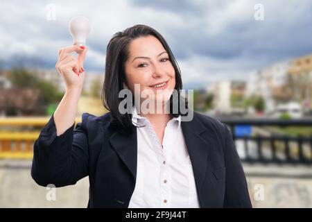 Glückliche Geschäftsfrau trägt Büroanzug halten Glühbirne als präsentiert gut Idee Konzept lächelnd auf Straße Natur Hintergrund Stockfoto