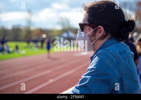 Ein junger Kaukasusmann in einem blauen Denim-Hemd trägt eine Sonnenbrille und eine Gesichtsmask, während er einem Leichtathletik-Event und einer laufenden Strecke zuschaut. Schließen Stockfoto