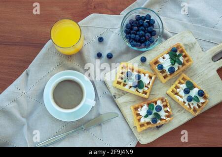 Waffeln mit Creme und Blaubeeren, eine Tasse Kaffee und Orangensaft auf dem Tisch auf einer Stoffserviette. Draufsicht, flach liegend. Stockfoto