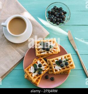 Tasse Kaffee und Waffeln mit Frischkäse und Heidelbeeren auf einem rosa Teller auf einem türkisfarbenen Tisch. Draufsicht, flach liegend. Stockfoto