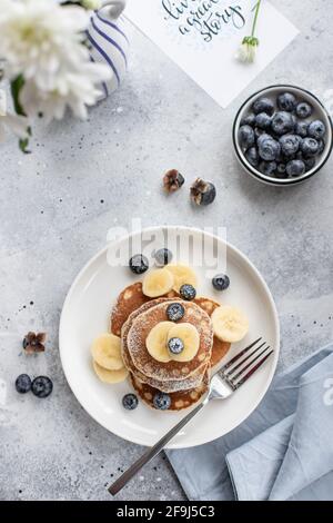 Leckere Pfannkuchen mit frischen Blaubeeren, Banane und Puderzucker auf grauem Betongrund. Vertikales Bild, Draufsicht, flach liegend Stockfoto