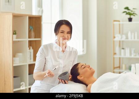 Lächelnder Dermatologe hält Glas mit feuchtigkeitsspendende Maske und bereit zu Tragen Sie es auf das Gesicht der Frau auf Stockfoto