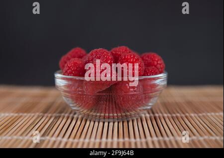 Zentriert, frisch gepflückte Himbeeren in einer Schale auf einer Bambusmatte vor dunklem Hintergrund, saftige, gesunde und saftige Früchte der Gattung Rubus Stockfoto