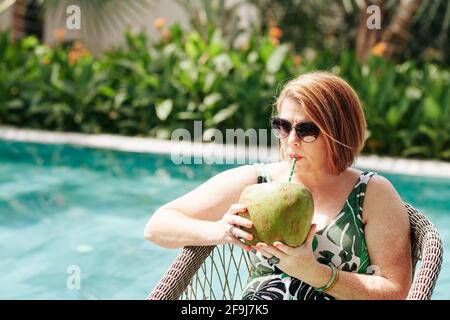 Reife Frau in Sonnenbrillen trinkt Kokosnusscocktails, wenn sie sich entspannt Die Sonne Stockfoto