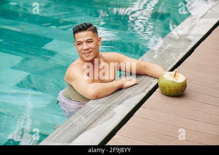 Fröhlicher Mann mittleren Alters, der im Swimmingpool des Wellnesshotels steht Und trinken süßen leckeren Kokosnusscocktail Stockfoto